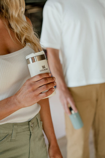A woman holding up a cream Stanley Aerolight Transit Mug alongside a man who is holding a Shale Stanley Aerolight Transit Mug