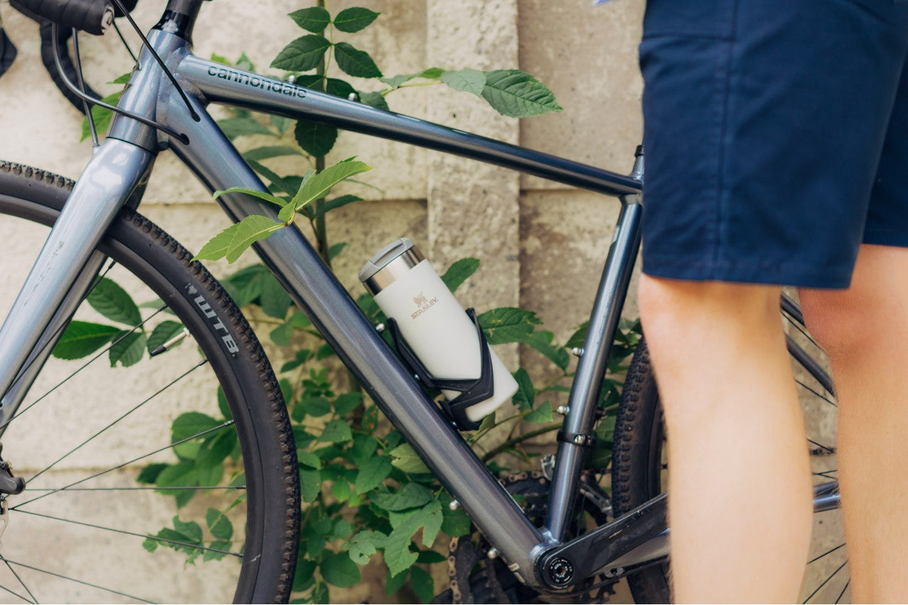 A mans legs shown next to a bicycle that has a cream Stanley Aerolight Transit Mug in the bicycle's bottle cage